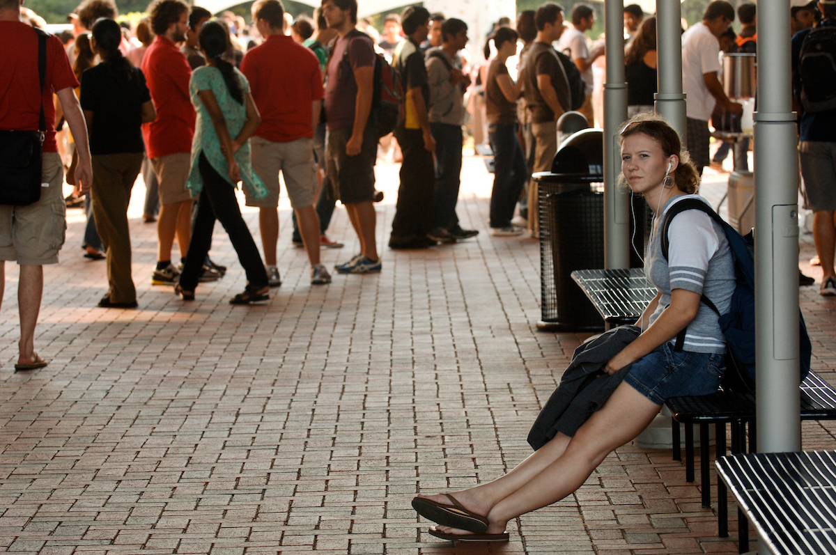 A female NC State student sits alone on a bench with a crowd in the background.